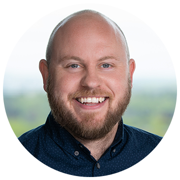 A man with a bright smile and a well-groomed beard is facing the camera. He wears a navy blue shirt and stands against a softly blurred outdoor background, suggesting a sunny day.