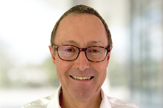 A man with short, dark hair and glasses smiles. He wears a white shirt. The background is blurred, suggesting an indoor environment with light colors and indistinct shapes.