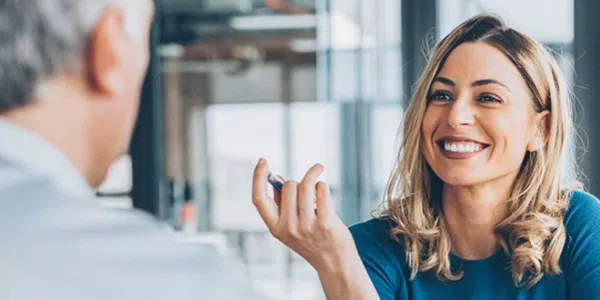 A smiling woman with wavy hair gestures while engaging in conversation across a table with a person whose back is visible. The setting is bright with large windows, suggesting a modern office environment.