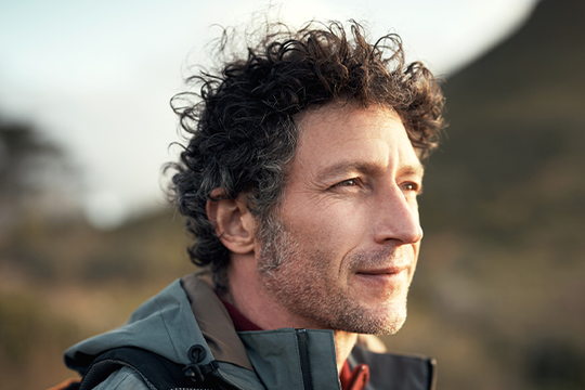 A man with curly hair gazes thoughtfully into the distance. He is dressed in outdoor clothing and stands against a blurred natural landscape, suggesting a setting of exploration or contemplation.
