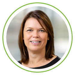 A woman with shoulder-length brown hair is smiling directly at the viewer. She is wearing a black top, and the background is softly blurred, suggesting an indoor environment.