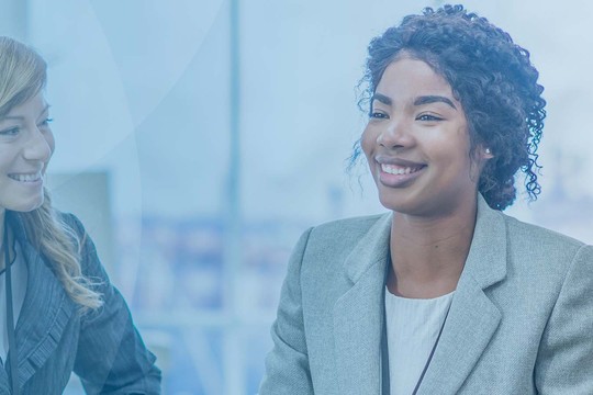 Three individuals are engaged in a conversation, smiling and sharing ideas, in a bright office setting with large windows and a light blue background.