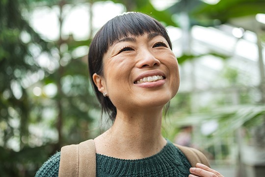 A woman with a backpack smiles brightly while looking upwards. She stands amidst lush greenery in a bright, airy environment, suggesting a greenhouse or botanical garden.