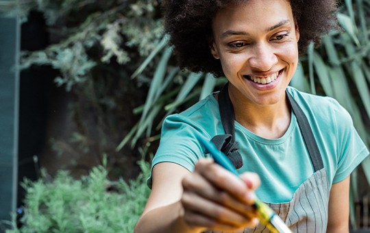 A woman with curly hair smiles while holding a tool, watering plants in a lush garden filled with green foliage, creating a vibrant, nurturing atmosphere.