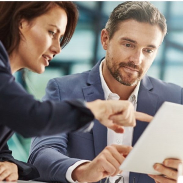 A woman points at a tablet held by a man, both engaged in discussion. They are seated at a table in a modern office setting with glass elements in the background.