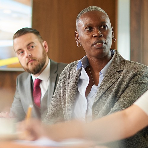 A professional woman talks attentively in a meeting room, while three colleagues listen. The atmosphere is focused and collaborative, with a background of wood paneling and a screen displaying information.