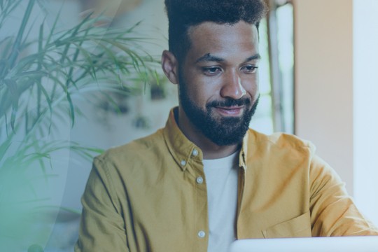 A man with a beard sits at a desk, smiling while using a laptop. He is surrounded by indoor plants and large windows that let in natural light.
