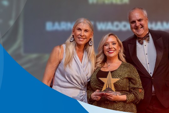 Three individuals stand together, smiling and holding an award in a festive setting, with a blue gradient background. The atmosphere is celebratory, likely at a formal event or ceremony.