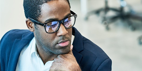 A man in glasses is thoughtfully resting his chin on his hand, wearing a navy blazer over a white shirt. The background features an office setting with blurred furniture.