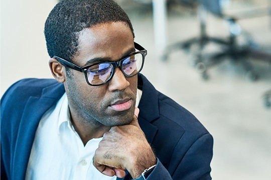 A man in glasses is thoughtfully resting his chin on his hand, wearing a navy blazer over a white shirt. The background features an office setting with blurred furniture.