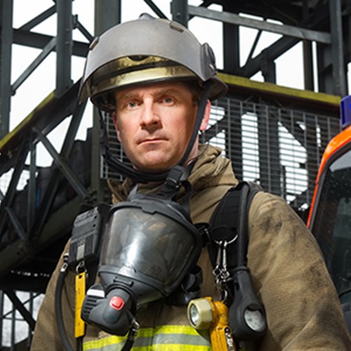A firefighter stands confidently in protective gear, wearing a helmet and a mask. In the background, a fire truck and a smoky building are visible, indicating an emergency response context.