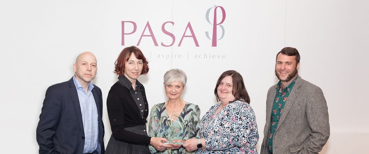 Five individuals stand together, with one holding an award. They are positioned against a white backdrop featuring the words "PASA" and "aspire | achieve," suggesting a formal recognition event.
