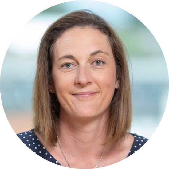 A woman with shoulder-length brown hair smiles gently. She wears a navy polka dot top and a necklace, set against a softly blurred outdoor background.