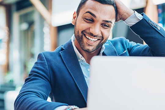 A man in a blue suit smiles while leaning towards a laptop, appearing engaged and happy. He is in a bright, modern indoor space, suggesting a relaxed atmosphere.