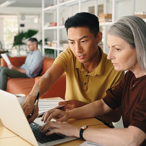 A young man and a middle-aged woman collaboratively work on a laptop at a table, sharing ideas, while a man sits in the background reading in a modern office space.