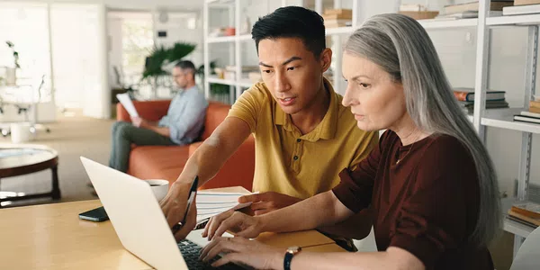 A young man and a middle-aged woman collaboratively work on a laptop at a table, sharing ideas, while a man sits in the background reading in a modern office space.