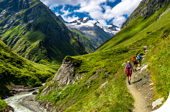 A rocky path winds through a lush green valley, where four hikers are walking alongside a river, surrounded by towering mountains and a bright blue sky with scattered clouds.