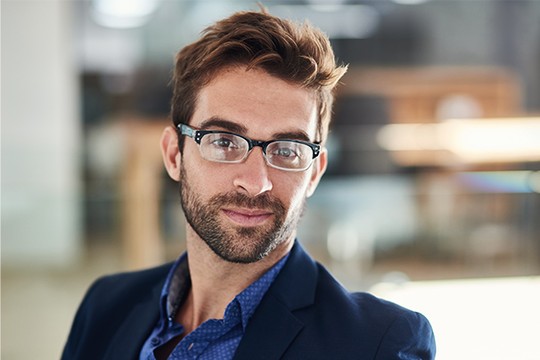 A man with glasses and a beard poses confidently, wearing a dark suit over a blue shirt. The background is softly blurred, suggesting a modern office environment.