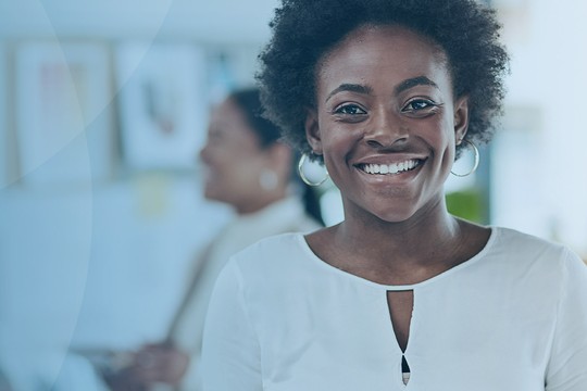 A smiling woman with curly hair stands confidently in a bright, modern workspace. People are engaged in conversation in the background, surrounded by plants and bookshelves.