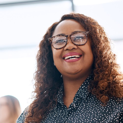 A woman with curly hair and glasses smiles brightly while standing in a bright, modern environment, possibly an office, engaging with others nearby.