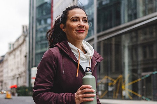A woman walks confidently on a street, holding a green water bottle. She wears a maroon hoodie layered over a white top, surrounded by modern buildings and a city atmosphere.