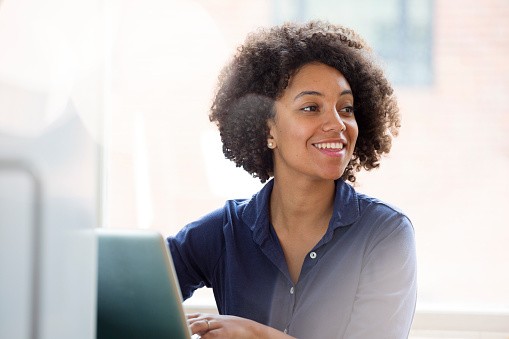 A young woman with curly hair smiles while sitting in front of a laptop, engaged in work or conversation. Natural light illuminates a bright, casual indoor space.