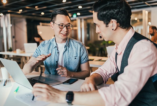 Two men are seated at a table, engaging in conversation while using a laptop. The setting is a modern workspace with other individuals in the background, indicating a collaborative environment.
