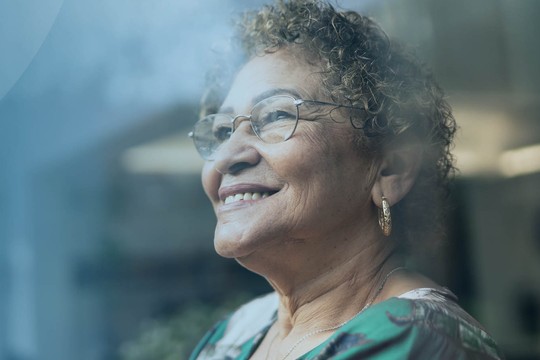 An older woman with curly hair smiles while gazing outside through a misted window, creating a reflective atmosphere. Soft blue hues dominate the background, adding warmth to the scene.