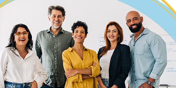 Five diverse professionals are standing together, smiling and posing. They are in a bright office setting, with whiteboards and colorful accents in the background.