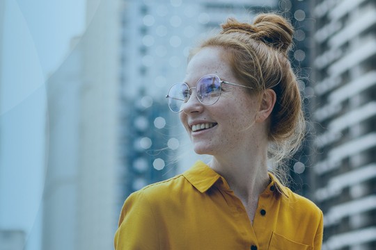 A woman with red hair in a yellow shirt smiles while looking to her side. She wears glasses, and modern buildings form a blurred background that suggests an urban setting.