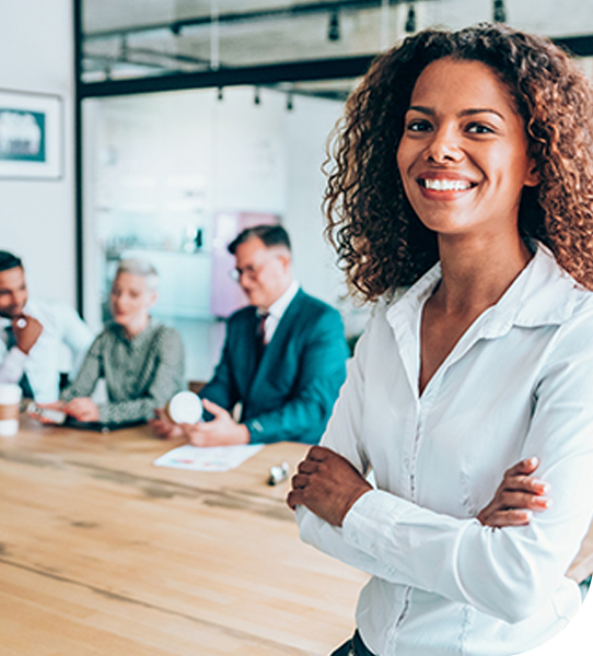 A smiling woman stands confidently in front of a wooden conference table. Behind her, three colleagues engage in discussion, contributing to a professional office environment with a modern aesthetic.