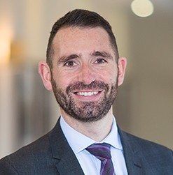 A man with a short beard smiles confidently, wearing a dark suit and patterned tie. He stands in a well-lit, modern environment with soft-focus background elements.