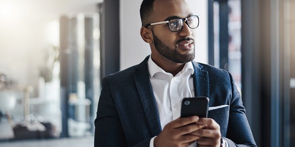A man in a suit stands indoors, holding a smartphone and engaged in conversation. He appears thoughtful, with large windows in the background allowing soft light to enter.