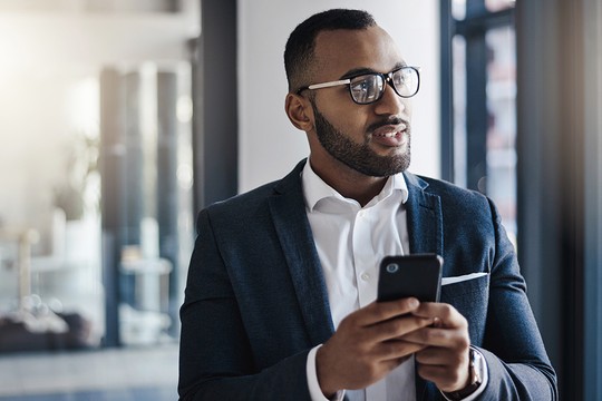 A man in a suit stands indoors, holding a smartphone and engaged in conversation. He appears thoughtful, with large windows in the background allowing soft light to enter.