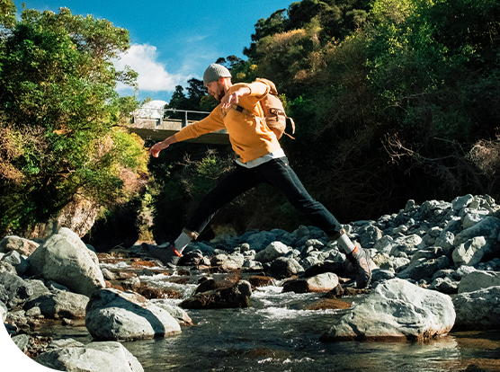 A person in a yellow sweater leaps across rocks in a shallow stream, surrounded by dense greenery and hills, with a blue sky and clouds visible in the background.