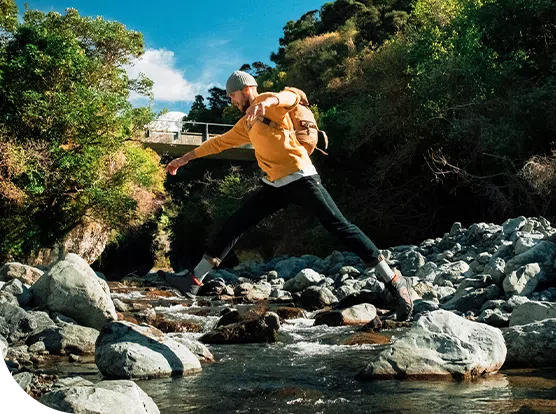 A person in a yellow sweater leaps across rocks in a shallow stream, surrounded by dense greenery and hills, with a blue sky and clouds visible in the background.