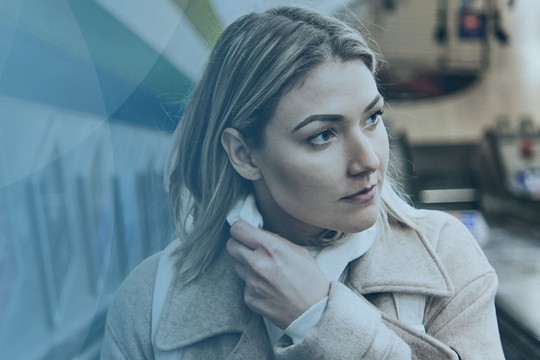 A woman with light brown hair is adjusting her collar while gazing thoughtfully. She is in a subway station, with an escalator in the background and people nearby.