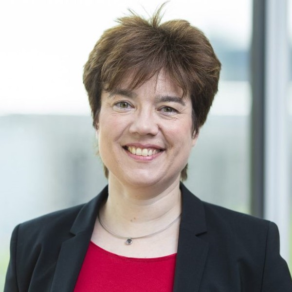 A smiling woman with short, dark hair wears a black blazer over a red top, standing indoors with a soft, blurred background of light windows.