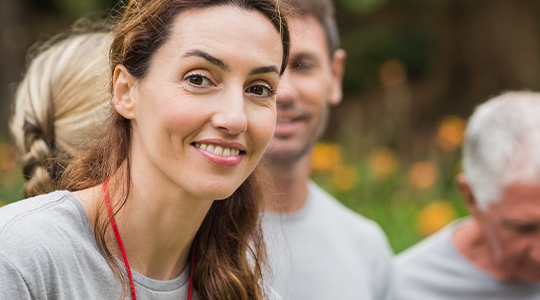 A woman smiles while participating in an outdoor activity, surrounded by a group of people in gray shirts, with a lush garden visible in the background.
