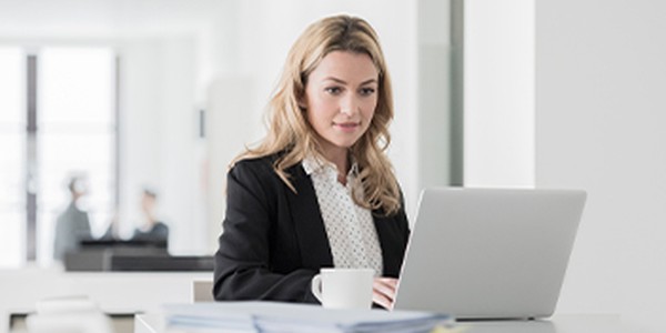 A woman in a black blazer is focused on her laptop while sitting at a modern desk. A cup is placed nearby, with a bright, minimalistic office setting in the background.
