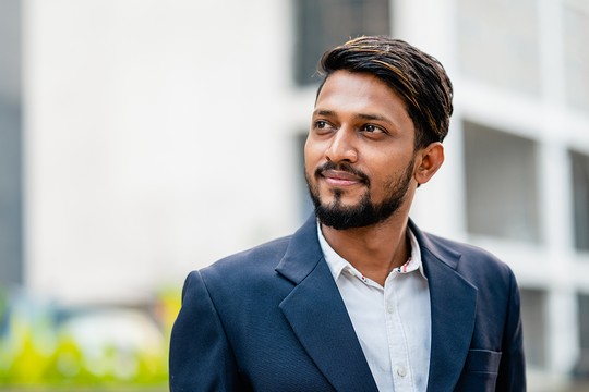 A man with a beard is smiling while looking slightly upward. He wears a dark blazer over a white shirt. The background features blurred buildings and greenery, suggesting an urban setting.