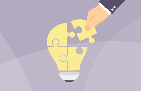 A hand is placing a yellow puzzle piece into a light bulb shape, symbolizing the completion of an idea or solution. The background is a soft, muted gray.