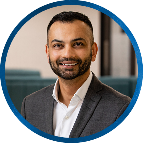A man in a suit smiles confidently while facing the camera. He is in a modern indoor setting with soft, neutral colors in the background, suggesting a professional atmosphere.