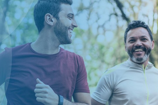 Two men are jogging together, smiling and enjoying their run in a lush green park. Sunlight filters through the trees, creating a vibrant, lively atmosphere.