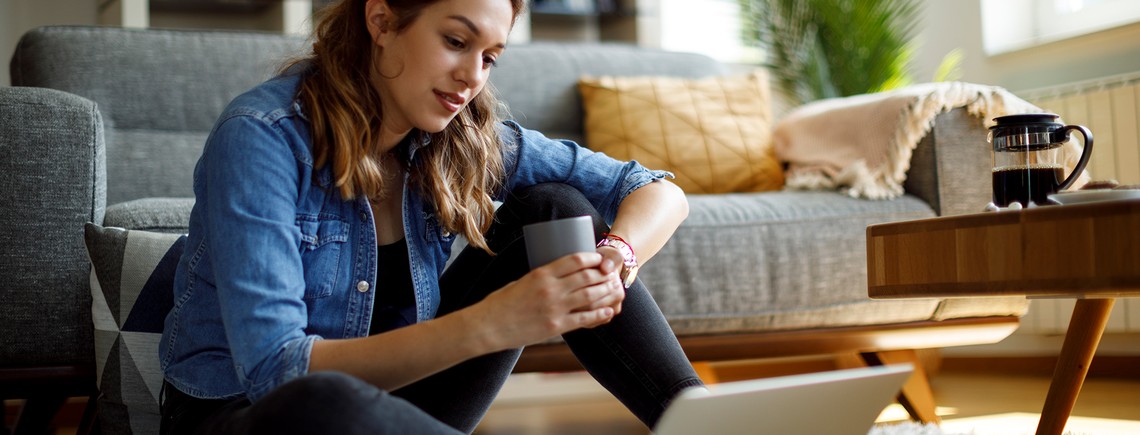 A woman sits on the floor, holding a mug and looking at a laptop. She is in a cozy living room with a sofa, coffee table, and a coffee press nearby.