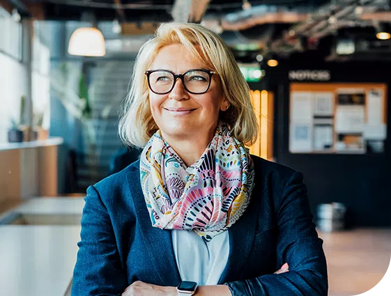 A woman stands confidently with her arms crossed, wearing glasses and a colorful scarf, in a modern indoor workspace filled with natural light, plants, and a dark wall displaying notices.