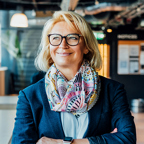 A woman stands confidently with her arms crossed, wearing glasses and a colorful scarf, in a modern indoor workspace filled with natural light, plants, and a dark wall displaying notices.