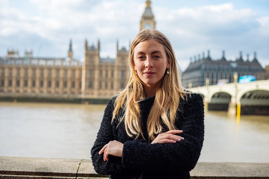 A woman with long blonde hair stands confidently, arms crossed, in front of a river with majestic architecture in the background, under a partly cloudy sky.