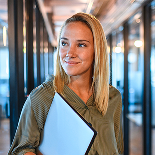 A woman with shoulder-length blonde hair holds a binder while smiling, walking through a modern office corridor illuminated by warm lighting and flanked by glass walls.