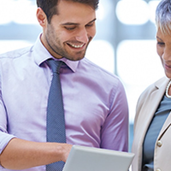 A young man and an older woman are discussing content on a tablet, smiling at each other. They are in a well-lit office environment, indicating a professional context.